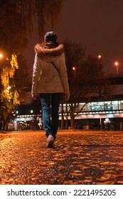 Woman Walks At Night In An Autumn Park. Rear View Of Young Female In White Coat Walking Away On Wet Alley With Orange Foliage