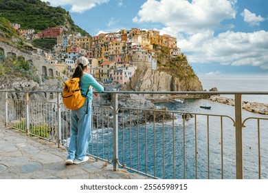 A woman wanderer stands by a railing, admiring the vibrant hues of the cliffside villages of Cinque Terre, as the serene sea sparkles under a cloudy sky, Manarola Italy during Autumn - Powered by Shutterstock