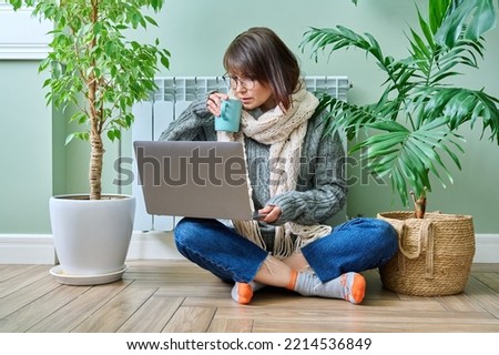 Woman in warm clothes sitting near heating radiator, using laptop