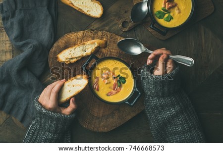 Woman in warm grey sweater eating corn creamy chowder soup with prawns served in individual pots, top view. Woman' s hand keeping spoon and bread slice. Flat-lay of rustic dinner table. Slow food