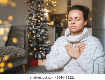 Woman in a warm knit sweater meditating and practicing breathing exercises at home during Christmas holidays, with a Christmas tree in the background. - Powered by Shutterstock