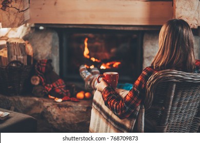 Woman In Warm Knitted Cardigan Sweater By Fireplace. Woman Relaxes By Warm Fire With A Cup Of Hot Drink And Warming Up Her Feet In Woollen Socks. Cozy Atmosphere. Winter And Christmas Holidays Concept