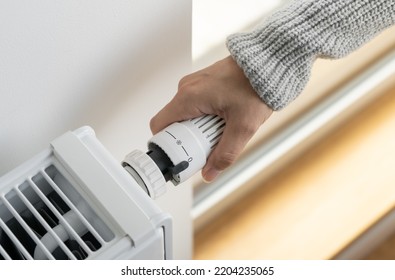 A Woman In A Warm Knitted Sweater Turns Off The Heating By Setting The Thermostat Valve Of The Radiator To The Minimum Value. Turning Off The Heating In The Apartment During The Energy Crisis.