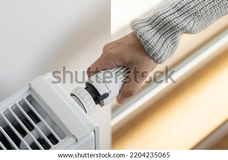 A woman in a warm knitted sweater turns off the heating by setting the thermostat valve of the radiator to the minimum value. Turning off the heating in the apartment during the energy crisis.