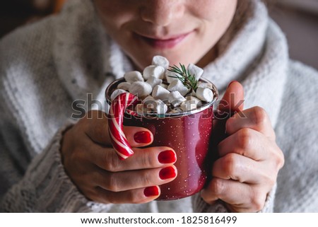 A woman in a warm sweater is enjoying of cup of hot chocolate decorated with marshmallows, red candy and a Christmas tree branch. Concept of a festive winter mood and cozy.