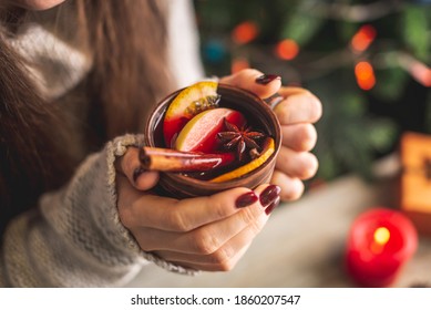 Woman in a warm sweater is holding in her hands a cup of aromatic hot mulled wine on the background of a Christmas tree with lights. Concept of a festive atmosphere and cozy winter mood. Closeup