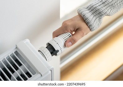 A woman in a warm sweater turns the regulator of the thermostatic radiator valve to the middle position to set the economical heating mode of the room. Heating of the apartment in the cold season. - Shutterstock ID 2207439695