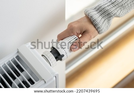 A woman in a warm sweater turns the regulator of the thermostatic radiator valve to the middle position to set the economical heating mode of the room. Heating of the apartment in the cold season. [[stock_photo]] © 