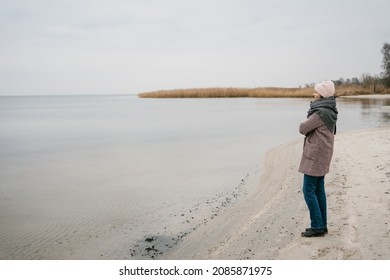 Woman in warm trendy winter outfit standing on a beach in winter on a cold bleak day staring thoughtfully out over a calm ocean with copyspace in a full length view - Shutterstock ID 2085871975