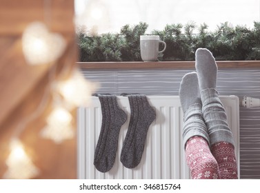 Woman Warming Up With Feet On Heater Winter Woolen Socks Drying On A Heater, Christmas Lights, Decorations And Hot Drink 