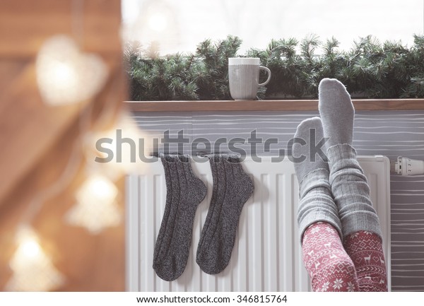 Woman warming up with feet on heater Winter woolen socks\
drying on a heater, christmas lights, decorations and hot drink\
