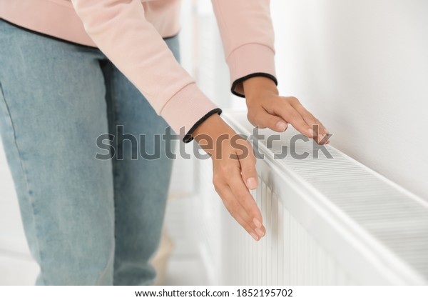 Woman warming hands on heating radiator near\
white wall, closeup
