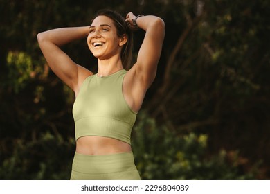 Woman warming up for a morning workout outdoors. Happy sports woman tying her hair and preparing for yoga. Fit woman standing against a nature background.