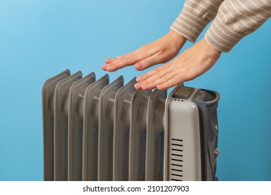 Woman warms her hands near hot oil filled electric heater against blue background. Using 9 fins space heater. Portable household appliance for heating home in a cold season. Front view.
