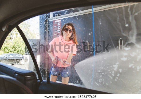 Woman washing car with pressure washer\
at self-service car wash station, view from the\
car