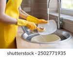 Woman Washing Dishes in Kitchen Sink During the Day, Wearing Yellow Gloves and Apron, Using Brush to Clean Plate