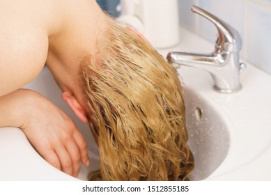 Woman washing her long blonde hair in bathroom sink. Haircare at home concept.