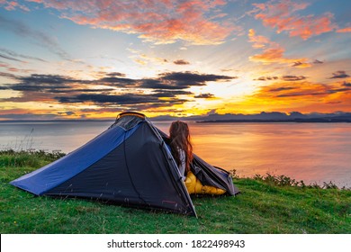 Woman Watching The Morning Sunrise Over Water From Her Tent While Wild Camping In The Isle Of Skye, Scotland, UK