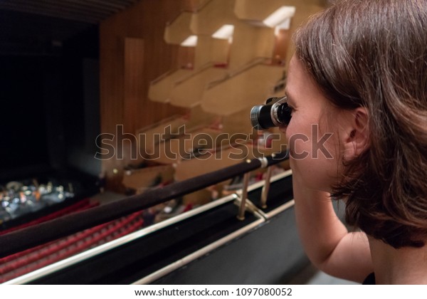 A woman is watching the orchestra in a theatre\
with opera glasses