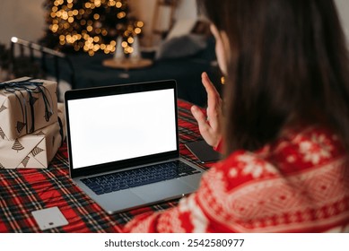 Woman waving to laptop screen during a video call with festive decorations, gifts, and Christmas lights, creating a cozy holiday ambiance. - Powered by Shutterstock