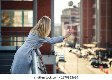 Woman Waving From Terrace To People At Street. Cityscape Background. Blond Hair Woman Wearing Stylish Grey Coat In City