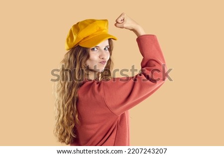 Woman with wavy hair in yellow cap beret and red sweatshirt demonstrates power. Optimistic active girl isolated on beige background looking at camera, smiling flexing arm with strong bicep muscle.