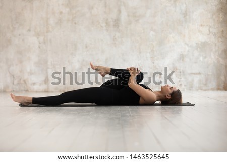 Woman wear black sport clothes lying on floor practising asana do Half Knees to Chest Pose near grunge wall beige textured background, help ease back pain, flexible body stretch for beginners concept