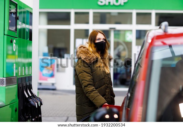Woman wear medical mask at self-service gas\
station, hold fuel nozzle, refuel the car with petrol during corona\
virus pandemic lockdown. People in masks as preventive measure and\
covid protection