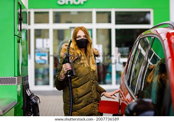 Woman wear medical mask at self-service gas
station, hold fuel nozzle, refuel the car with petrol during corona
virus pandemic lockdown. People in masks as preventive measure and
covid protection