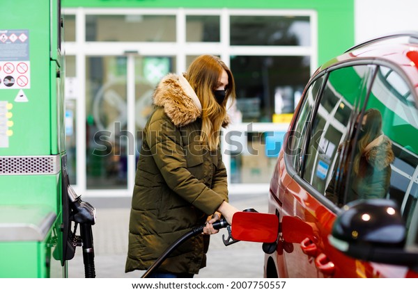 Woman wear medical mask at self-service gas\
station, hold fuel nozzle, refuel the car with petrol during corona\
virus pandemic lockdown. People in masks as preventive measure and\
covid protection