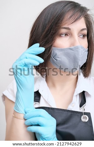 woman wearing apron putting on blue protective gloves. brunette woman with textile mask on a face getting ready to do beauty procedure. depilation beautician