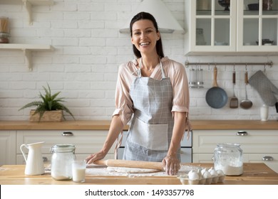 Woman wearing apron smiling looking at camera standing in kitchen preparing for family dinner holding rolling pin flattening dough eggs flour milk on wooden table, housewife chores or hobby concept