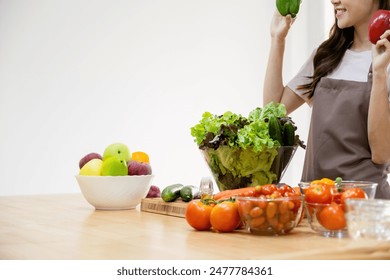  Woman wearing apron standing at counter with healthy ingredients, vegetables, and fruits in the kitchen at home holding bell peppers to pick and choose for cooking healthy meal.  - Powered by Shutterstock