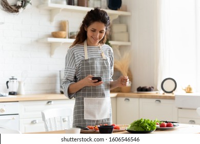 Woman Wearing Apron Standing On Domestic Kitchen Table Full Of Fresh Vegetables, Housewife Holds Phone Using Cooking Apps Websites Search Recipes, Chatting With Friend Distracted From Food Preparation