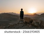 Woman wearing backpack stands alone on the high rock in the dark during sunset and looking at setting sun in the desert mountains of Negev desert in Israel