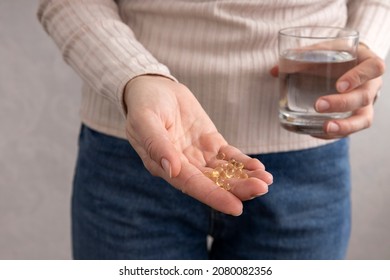 Woman Wearing Beige Sweater And Jeans At Home, Holding Vitamin D Capsules And Glass Of Water