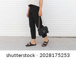 Woman wearing beige t-shirt, black pants, bag and flat sandals walking outdoor near white roller door. Details of stylish trendy basic minimalistic casual outfit. Street fashion. Women