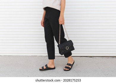 Mujer con camiseta beige, pantalones negros, bolsa y sandalias planas caminando al aire libre cerca de la puerta de un rodillo blanco. Los detalles de diseño minimalista básico y casual. Moda callejera. piernas de las mujeres, sin cara