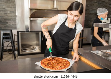 Woman Wearing Black Apron Using Pizza Cutter To Cut Slices Of Freshly Baked Pepperoni Salami Pizza In Pizza Shop