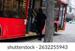 A woman wearing black clothing approaches a bright red bus at a city bus stop. The trees and pavement indicate a typical urban setting early in the day.
