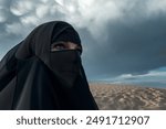 A woman wearing a black niqab looks towards a dramatic stormy sky over desert dunes. Articles on Muslim culture, faith, women