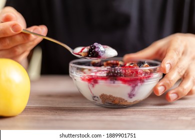 Woman Wearing Black Shirt Eats A Fresh Homemade Glass Bowl Of Creamy Yogurt Parfait With Berries, Muesli And Seeds In It On Wooden Table Closeup Photo Shows Hand Holding A Spoon With An Apple On Side.