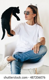 Женщина в Blanc футболка, джинсы и кроссовки, сидя на стуле и играя с черным котенком, тонированное фото, передняя футболка макет на модели, хипстерский стиль