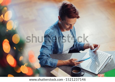 Woman wearing blue denim shirt sitting at the table by the Christmas tree with a credit card in her hand and shopping online on smartphone