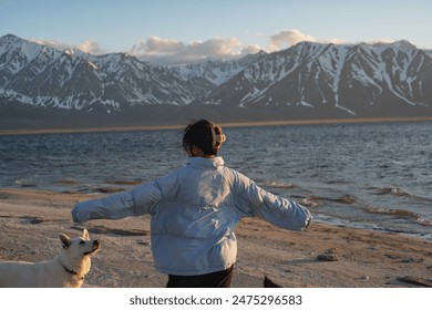 A woman wearing a blue jacket stands on a sandy beach with her white dog, looking out towards a snow-capped mountain range at sunset. - Powered by Shutterstock