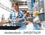 A woman wearing a blue and white plaid shirt is working on an assembly line. She is holding a circuit board in her hands. There are many bins and other electronic parts on the table in front of her.