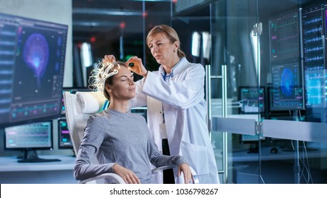 Woman Wearing Brainwave Scanning Headset Sits in a Chair while Scientist Adjusts the Device, Looks at Displays. In the Modern Brain Study Laboratory Monitors Show EEG Reading and Brain Model.