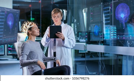 Woman Wearing Brainwave Scanning Headset Sits in a Chair while Scientist Adjusts the Device, Uses Tablet Computer. In the Modern Brain Study Laboratory Monitors Show EEG Reading and Brain Model.