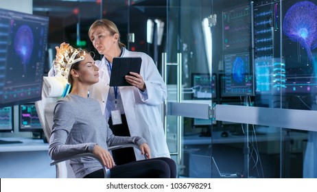 Woman Wearing Brainwave Scanning Headset Sits in a Chair while Scientist Adjusts the Device, Uses Tablet Computer. In the Modern Brain Study Laboratory Monitors Show EEG Reading and Brain Model.