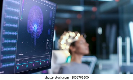 Woman Wearing Brainwave Scanning Headset Sits In A Chair In The Modern Brain Study Laboratory/ Neurological Research Center. Monitors Show EEG Reading And Brain Model.
